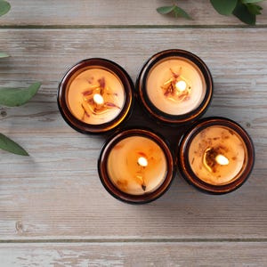May include: Four amber glass candle jars with a light brown wax and a flickering flame in each. The candles are arranged in a circle on a light brown wooden surface.