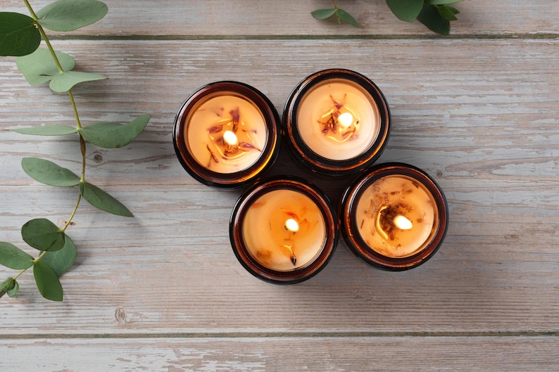 May include: Four amber glass candle jars with a light brown wax and a flickering flame in each. The candles are arranged in a circle on a light brown wooden surface.