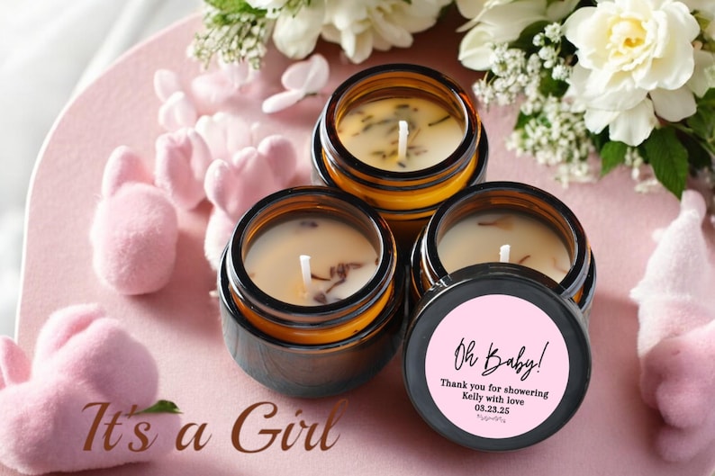 May include: Three small, round candles in amber-colored glass jars with black lids. The candles have white wax and wicks, with dried botanicals. One lid has a pink label with the text "Oh Baby!" and a message. The phrase "It's a Girl" is also visible.