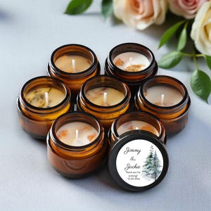 May include: Seven small, round, amber glass jars filled with wax candles. The candles have white wicks and decorative elements. One jar has a black lid with the text "Jimmy & Jackie" and a winter tree design.