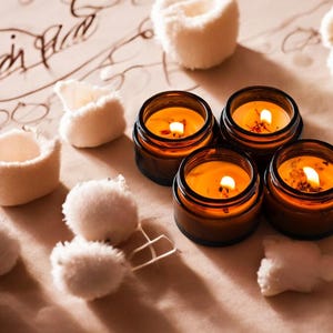 May include: Four amber glass jars containing lit candles with orange wax, each featuring a visible wick and flame. The candles are arranged in a cluster alongside several small, fluffy, white pom-poms on a light tan surface with dark script text.