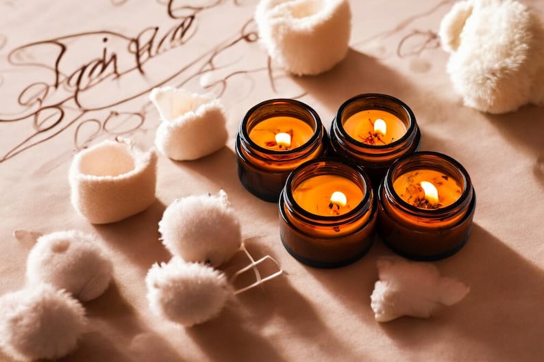 May include: Four amber glass jars containing lit candles with orange wax, each featuring a visible wick and flame. The candles are arranged in a cluster alongside several small, fluffy, white pom-poms on a light tan surface with dark script text.