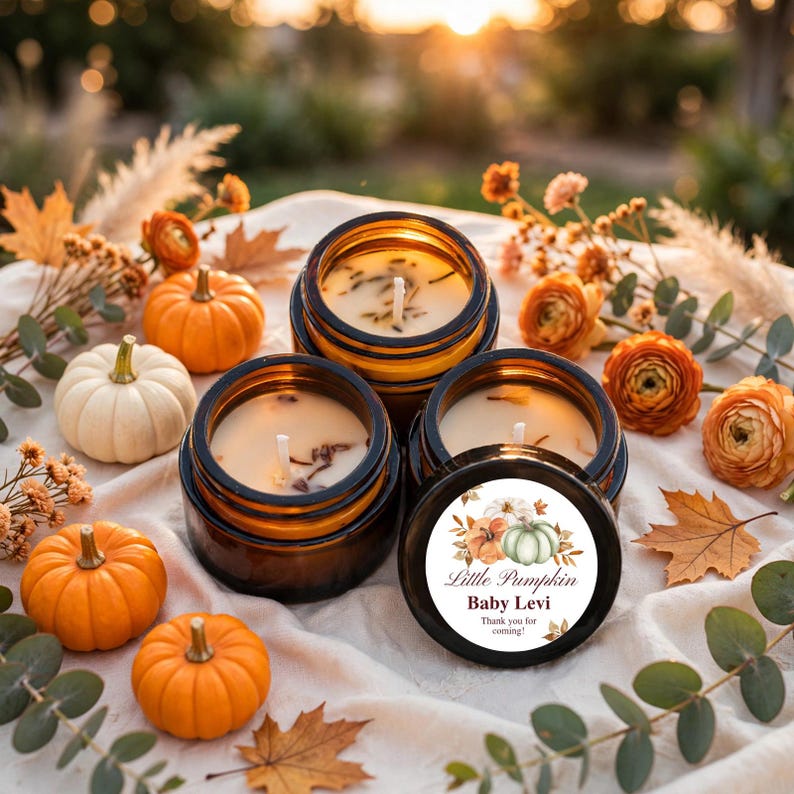 May include: Three amber glass jars containing cream-colored wax candles with visible wicks. One lid features a circular label with a watercolor pumpkin illustration and the text "Little Pumpkin Baby Levi Thank you for coming!" surrounded by autumn decor.