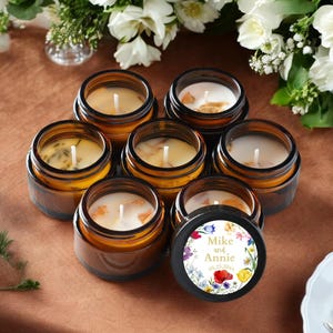 May include: A collection of seven amber glass jars containing white wax candles with single wicks. One jar lid features a circular label with a colorful floral border and the printed text Mike and Annie 05.25.20xx in a gold serif font.
