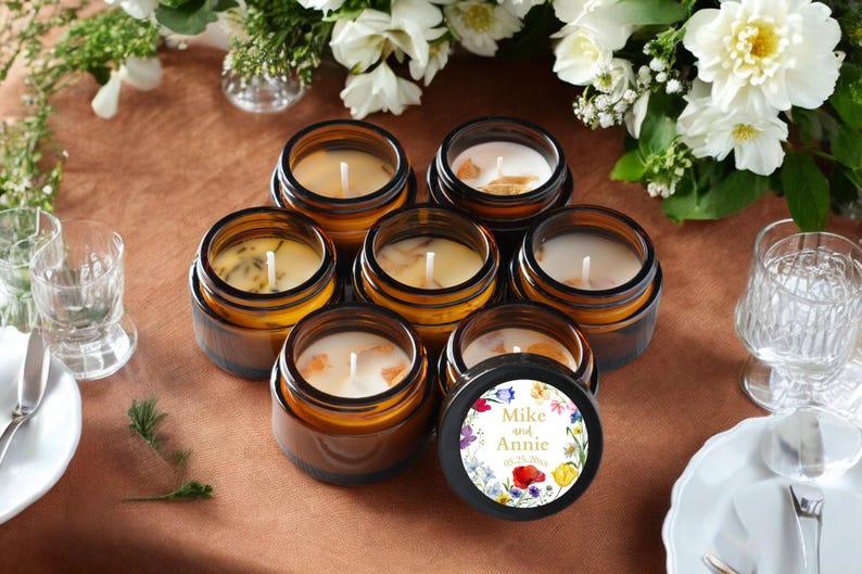 May include: A collection of seven amber glass jars containing white wax candles with single wicks. One jar lid features a circular label with a colorful floral border and the printed text Mike and Annie 05.25.20xx in a gold serif font.