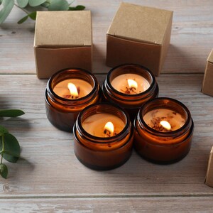 May include: Four amber glass candle jars with lit wicks, sitting on a light brown wooden surface. The candles are surrounded by four brown cardboard boxes.