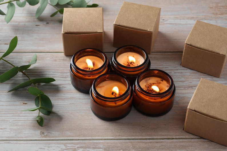 May include: Four amber glass candle jars with lit wicks, sitting on a light brown wooden surface. The candles are surrounded by four brown cardboard boxes.