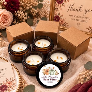 May include: Four amber glass jars filled with white wax candles, each featuring a single wick and small orange accents. One black lid displays a circular label with watercolor pumpkins and the text Little Pumpkin Baby Perry 11.02.24. Three brown cardboard boxes accompany the candles.