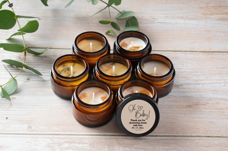 May include: Seven small, round, amber-colored glass jars filled with white wax candles. Some candles have decorative elements. One jar has a black lid with the text "Oh Baby!" and a message. Green leaves are in the background.