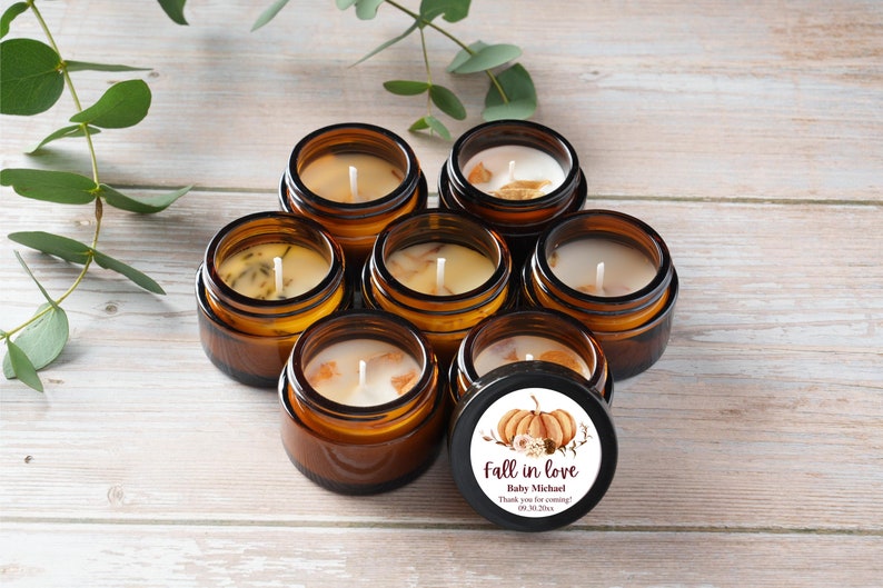 May include: A collection of small amber glass jars with white candles inside. The jars are arranged in a circle on a light wood surface. A single jar has a round sticker with a pumpkin design and the text "Fall in love Baby Michael Thank you for coming!"