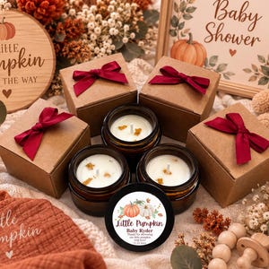 May include: Three amber glass candle jars with white wax and wicks, accompanied by three matching brown cardboard gift boxes tied with deep red satin ribbons. One black candle lid features a pumpkin graphic and the text Little Pumpkin Baby Ryder.