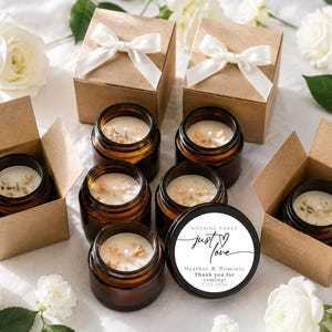 May include: A collection of amber glass candle jars with white wax, some placed inside open brown cardboard boxes with white ribbon bows. One black lid features the text: Nothing fancy just love, Heather & Dominic, Thank you for coming, 7.25.2026.