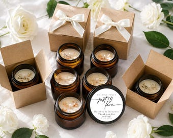 May include: A collection of amber glass candle jars with white wax, some placed inside open brown cardboard boxes with white ribbon bows. One black lid features the text: Nothing fancy just love, Heather & Dominic, Thank you for coming, 7.25.2026.