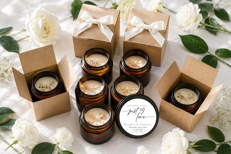 May include: A collection of amber glass candle jars with white wax, some placed inside open brown cardboard boxes with white ribbon bows. One black lid features the text: Nothing fancy just love, Heather & Dominic, Thank you for coming, 7.25.2026.