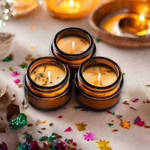 May include: Three small amber glass jars containing cream-colored wax candles with visible wicks and embedded botanical pieces. The candles are arranged in a cluster, each featuring a central white wick surrounded by light-colored wax.