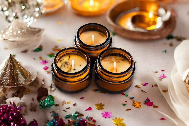 May include: Three small amber glass jars containing cream-colored wax candles with visible wicks and embedded botanical pieces. The candles are arranged in a cluster, each featuring a central white wick surrounded by light-colored wax.