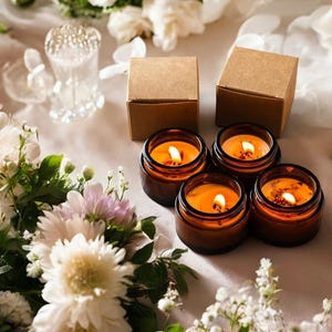 May include: Four amber glass jars containing lit candles with visible flames, arranged alongside four small square brown cardboard boxes. The scene is decorated with white and light pink flowers, including chrysanthemums and small blossoms.
