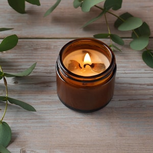 May include: A brown glass candle jar with a lit candle inside. The candle is surrounded by green eucalyptus leaves on a light wood surface.