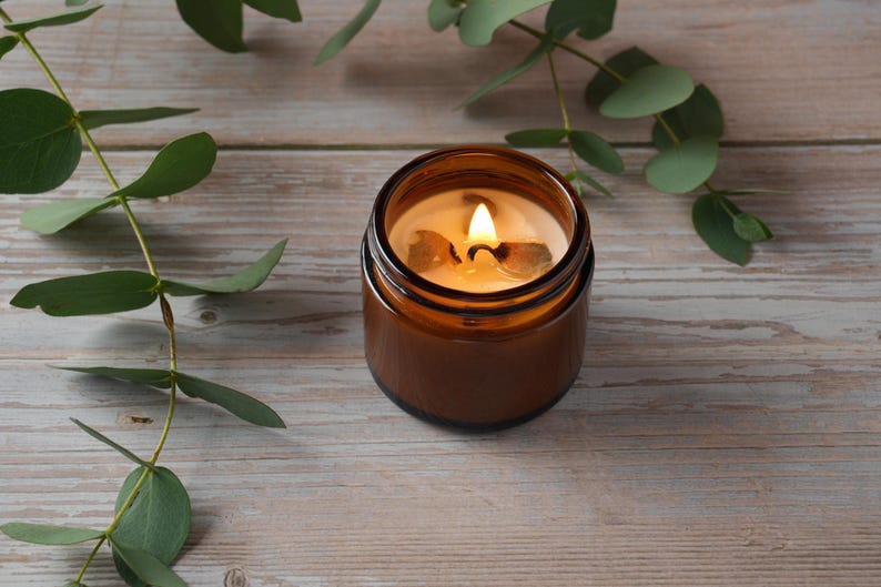 May include: A brown glass candle jar with a lit candle inside. The candle is surrounded by green eucalyptus leaves on a light wood surface.
