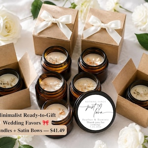 May include: Several amber glass candle jars filled with white wax, some placed inside open brown cardboard boxes with white satin bows. One black candle lid features the text: Nothing Fancy, just love, Heather & Dominic, Thank you for coming, 7.25.2026.