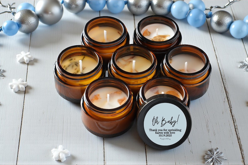 May include: A collection of small, amber-colored glass jar candles with white wax and wicks. One candle has a black lid with the text "Oh Baby!" and a personalized message. The candles are arranged on a white wooden surface.