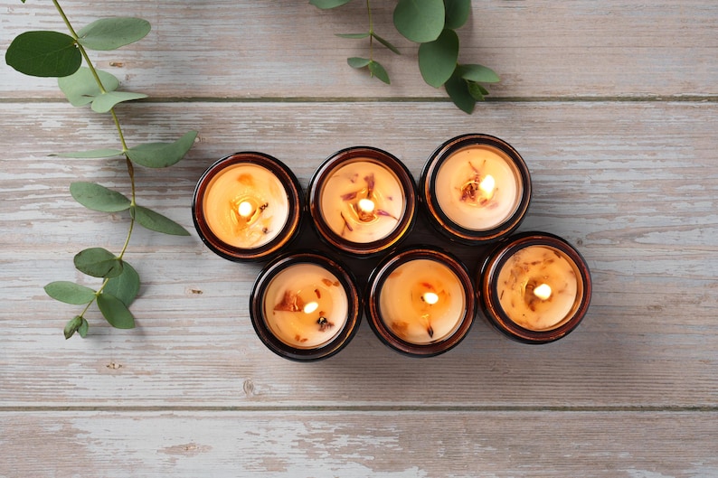 May include: Six amber glass candle jars with a natural wax blend and a warm, golden glow. The candles are arranged in a semi-circle on a light wood surface.