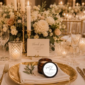 May include: A small amber glass candle jar with a round white label featuring the text "just love", "Heather & Dominic", and "Thank you for coming! 7.12.2025". The candle sits on a gold charger plate with a white napkin and a small green sprig.