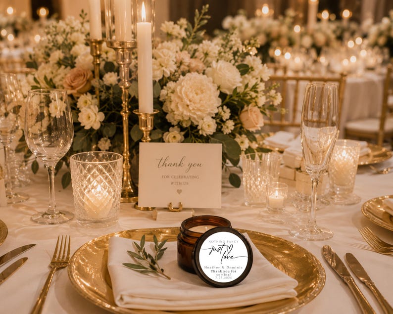 May include: A small amber glass candle jar with a round white label featuring the text "just love", "Heather & Dominic", and "Thank you for coming! 7.12.2025". The candle sits on a gold charger plate with a white napkin and a small green sprig.