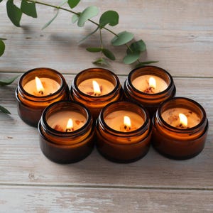 May include: Six amber glass candle jars with lids, each containing a lit candle with a warm, yellow glow. The candles are arranged in a semi-circle on a light wood surface.