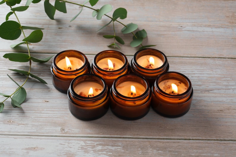 May include: Six amber glass candle jars with lids, each containing a lit candle with a warm, yellow glow. The candles are arranged in a semi-circle on a light wood surface.