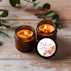 May include: Two amber glass jar candles with lit wicks. One candle has a black lid featuring a circular white label with pink text that reads "thank you! Emily's BRIDAL BRUNCH 04.23.20XX" and a colorful floral illustration with pink, orange, and white flowers.