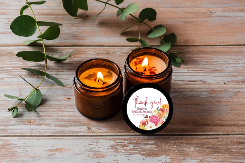 May include: Two amber glass jar candles with lit wicks. One candle has a black lid featuring a circular white label with pink text that reads "thank you! Emily's BRIDAL BRUNCH 04.23.20XX" and a colorful floral illustration with pink, orange, and white flowers.