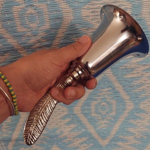 Op de afbeelding: Een zilveren bel met een handvat in de vorm van een veer. De bel wordt in een hand gehouden tegen een blauwe en witte gepatroneerde achtergrond.