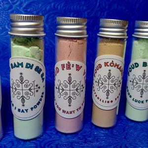 Può includere: Sei fiale di vetro riempite di polveri colorate, ciascuna sigillata con un tappo argentato. Le fiale hanno etichette con testo e un emblema decorativo. Le polveri sono di vari colori, tra cui rosa, verde e beige. Le fiale sono su una superficie blu.