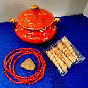 May include: A red and gold ceramic pot with a lid, a red beaded necklace, a small brown stone, and a bag of seashells on a blue surface.