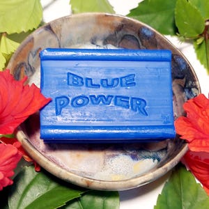 Puede incluir: Una pastilla de jabón azul con las palabras "BLUE POWER" en relieve. El jabón está en un pequeño plato de cerámica rodeado de flores de hibisco rojas y hojas verdes.