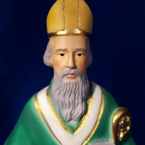 Puede incluir: Una figurita de un hombre que lleva una túnica verde y una mitra dorada y amarilla. Tiene barba blanca y sostiene un libro en su mano izquierda.