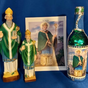 May include: Three St. Patrick figurines, one large and two small, with green and gold robes, holding a book and a staff. A green and blue bottle with a St. Patrick image and glitter is also shown. The bottle has a blue and gold label with the text "St. Patrick".