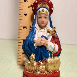 May include: A small, blue and red figurine of the Virgin Mary. She is wearing a blue robe and a crown. She is holding a sword and a rosary. The base of the figurine is red and has the words "Metrestilla" and "Cabe" written on it.