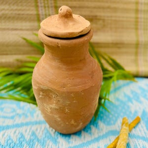 May include: A small, handcrafted terracotta jar with a lid. The jar is a warm, earthy brown color and has a rounded body with a slightly flared rim. The lid has a small knob on top. The jar is sitting on a blue and white patterned surface.
