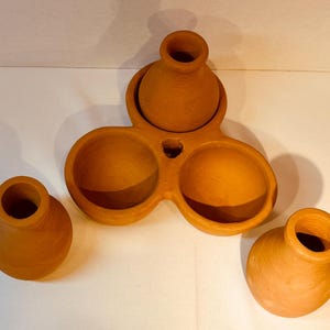 May include: A set of terracotta pottery. The set includes three small, round pots and a larger, three-sectioned serving dish. The pottery has a warm, earthy tone and a simple, rustic design.