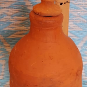May include: A terracotta clay bottle with a lid. The bottle is a light brown colour and has a smooth, slightly textured surface. The lid is a darker brown colour and has a rounded top.