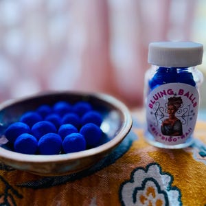 May include: A small glass jar filled with blue balls, labeled "BLUING BALLS" with a graphic of a woman. A ceramic bowl with blue balls sits next to the jar on a patterned surface.