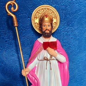 May include: A figurine of a saint holding a book and a staff. The saint is wearing a pink robe with gold trim and a gold mitre. The background is a blue textured surface.