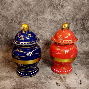 May include: Two decorative ceramic jars with lids. The jar on the left is blue with gold accents and white dots. The jar on the right is red with gold accents and white dots.