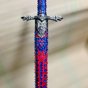 May include: A decorative pen with a sword design. The pen features a pink barrel adorned with red and blue rhinestones. The hilt and pommel are silver-toned, with intricate detailing. The pen's top is also embellished with blue rhinestones.