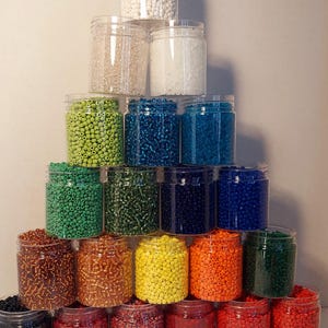 May include: A pyramid of clear plastic jars filled with colorful beads. The beads are in various colors, including white, green, blue, brown, yellow, orange, and red. The jars are stacked on top of each other.