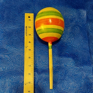 May include: A wooden toy with a round, yellow, green, and orange striped head on a stick. The toy is about 6 inches long.