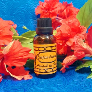May include: A small amber glass bottle with a black cap and a yellow label featuring the text "Parfum d'amour". The bottle is surrounded by vibrant red hibiscus flowers and green leaves against a blue background.