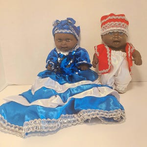 May include: Two dolls dressed in traditional attire. One doll wears a blue dress with white accents and a matching head wrap. The other doll is dressed in white pants, a red vest, and a white and red hat. Both dolls have dark skin and are on a white background.